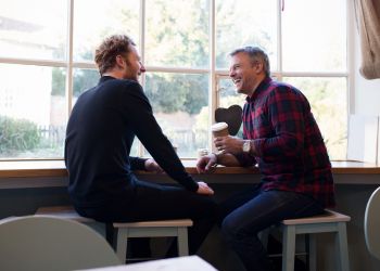 Attain Downtown apartment homes with Two men sit by a window in a café, laughing and chatting while one holds a coffee cup.