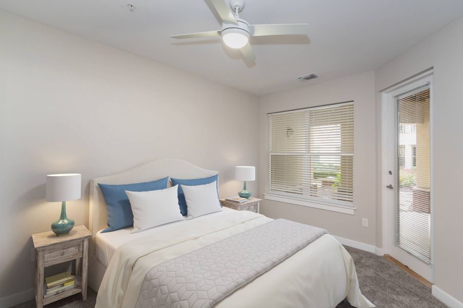 Attain Downtown East apartment homes with Bright bedroom with a bed, two side tables, lamps, a ceiling fan, and window blinds overlooking a patio.