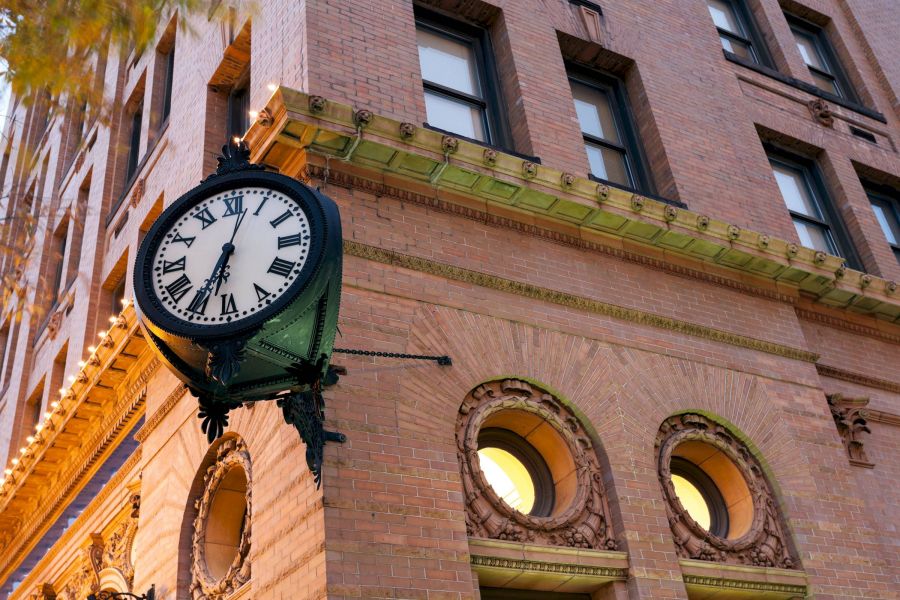 Attain Downtown apartment homes with A vintage clock showing 4:48 is mounted on the corner of a brick building with arched windows.