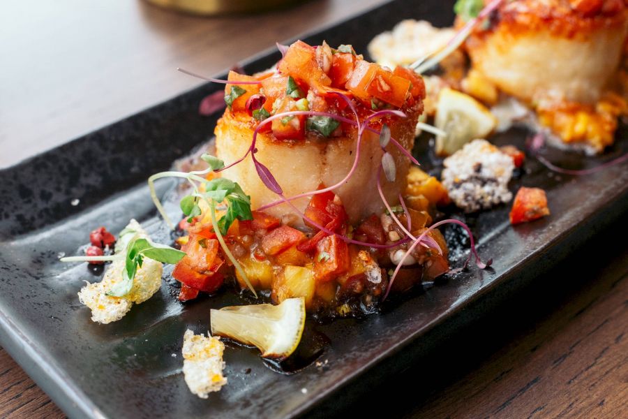 Attain Downtown apartment homes with Seared scallops topped with diced vegetables and microgreens, served on a black rectangular plate with lemon wedges.