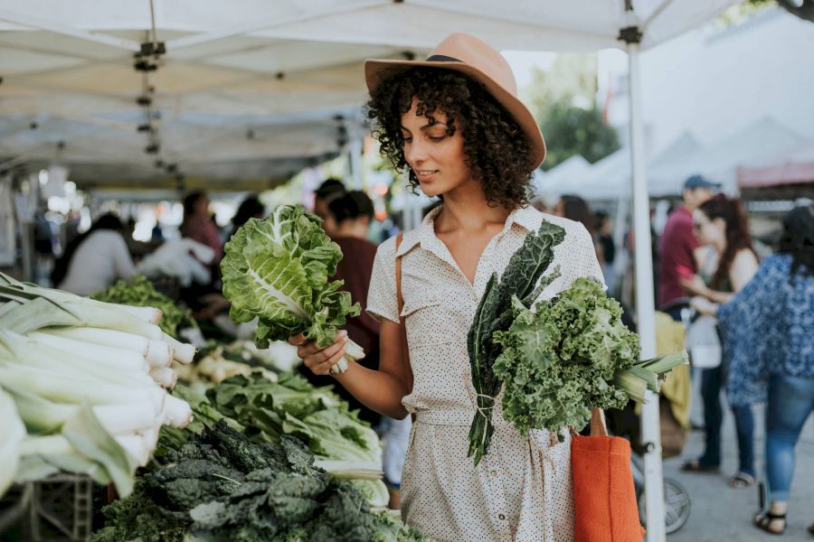 Attain Downtown apartment homes with Woman in a hat shopping for leafy greens at an outdoor farmers market, holding lettuce and kale.