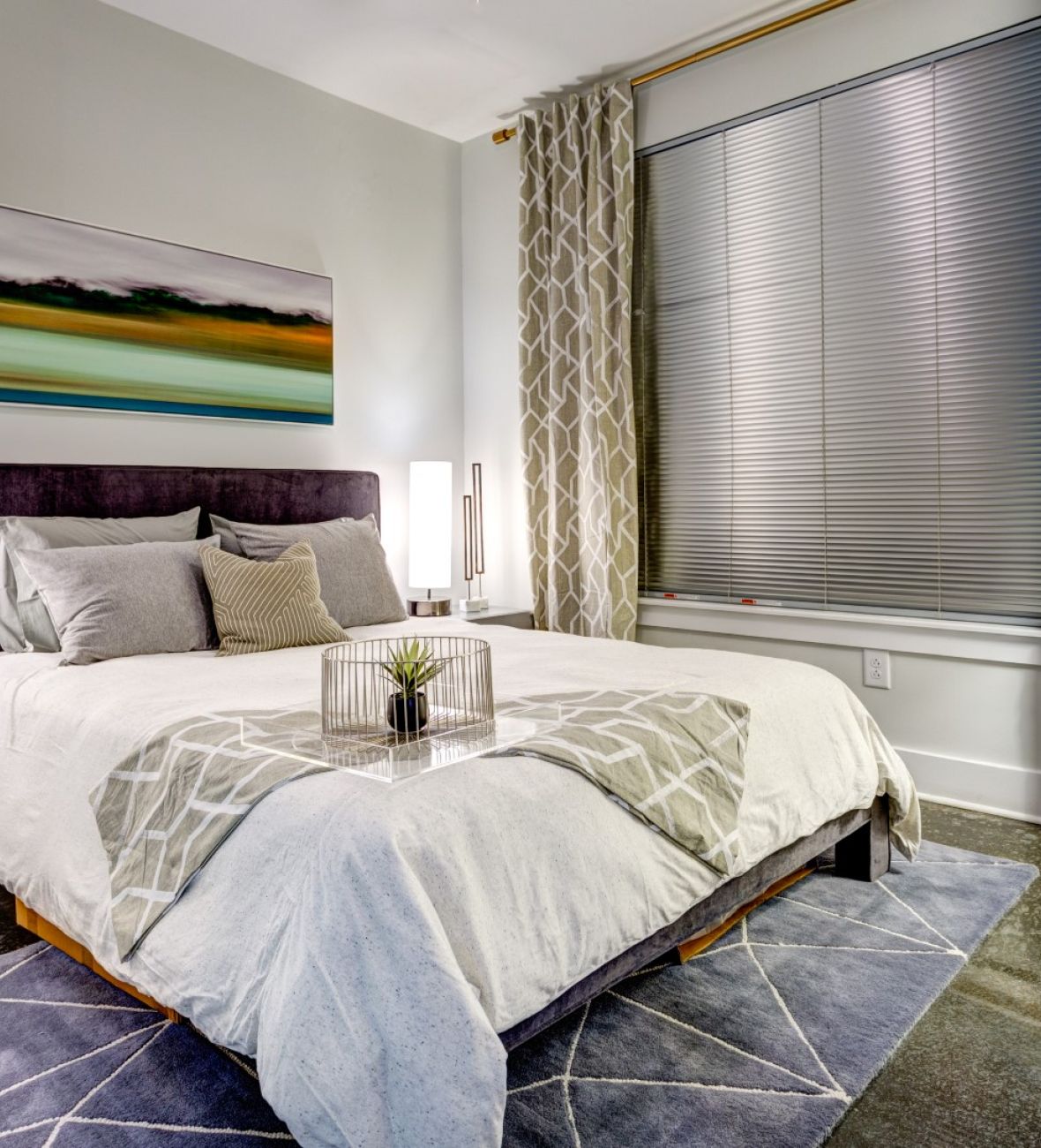 Attain Downtown apartment homes with Modern bedroom with a large bed, bedside tables, lamps, abstract art, and patterned curtains.