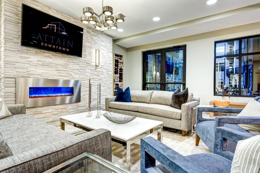 Attain Downtown apartment homes with Modern lounge with grey sofas, blue armchairs, a wall-mounted TV, electric fireplace, and large windows.