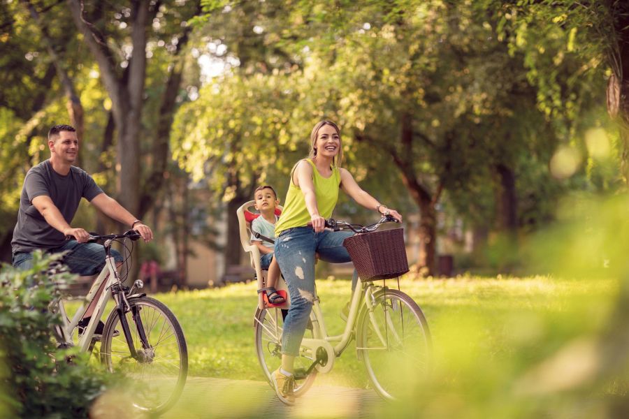 Attain on 5th apartment homes with A smiling family rides bicycles through a sunny, green park; a child sits on the mother's bike.