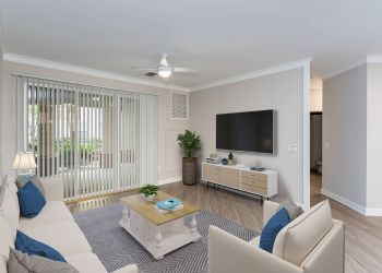 Attain Downtown East apartment homes with Modern living room with beige sofas, blue accents, TV on the wall, and large windows with vertical blinds.