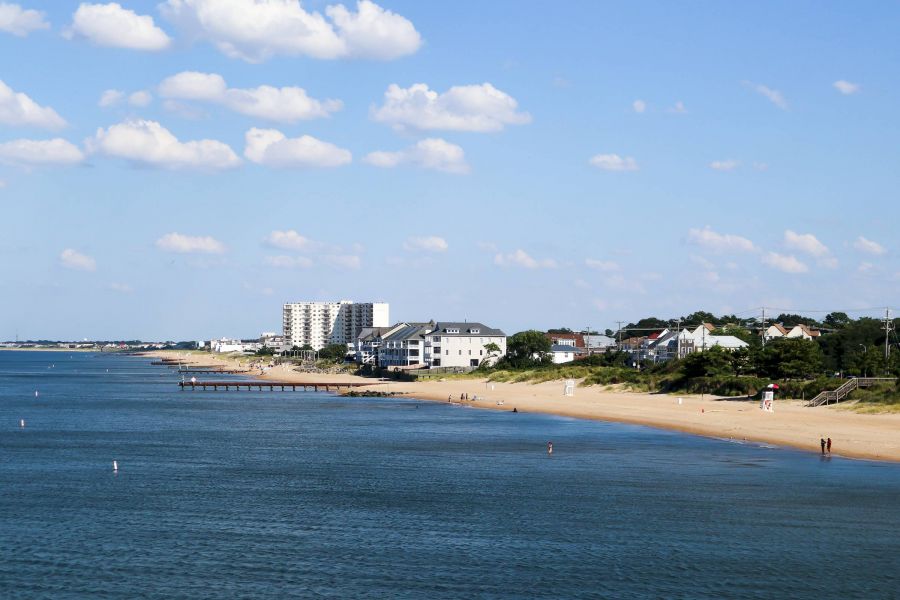 Attain Downtown apartment homes with Sandy beach with calm blue water, a pier, and buildings along the shoreline under a partly cloudy sky.