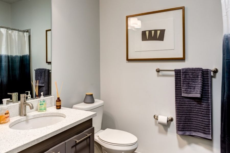Attain Downtown apartment homes with Modern bathroom with gray vanity, toilet, wall art, purple towels, and bright lighting.